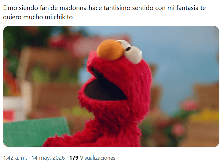 A close-up of a red puppet character with orange eyes and a smiling mouth, expressing excitement, set in a colorful background with greenery and vibrant decorations.